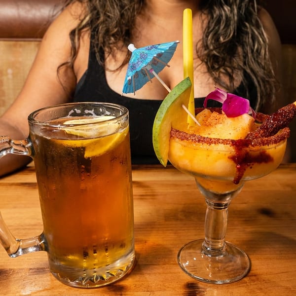 A person with a beer mug and a mango margarita with a blue umbrella and mango slice.