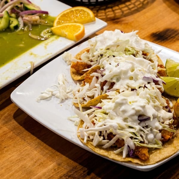 Two fish tacos with cabbage and crema, ceviche, and lime wedges on a wooden table.