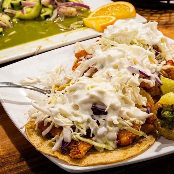 Two fish tacos topped with cabbage and white sauce, with a side of lime and a roasted pepper.
