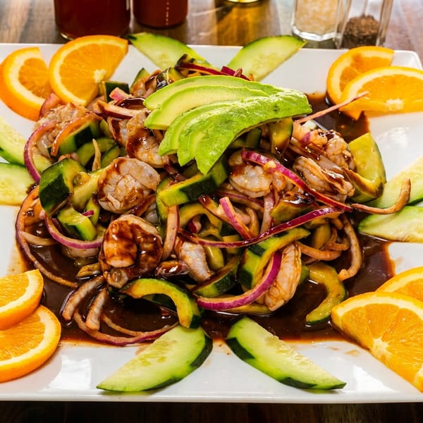 Shrimp ceviche with cucumber, red onion, avocado, and orange slices on a white plate.