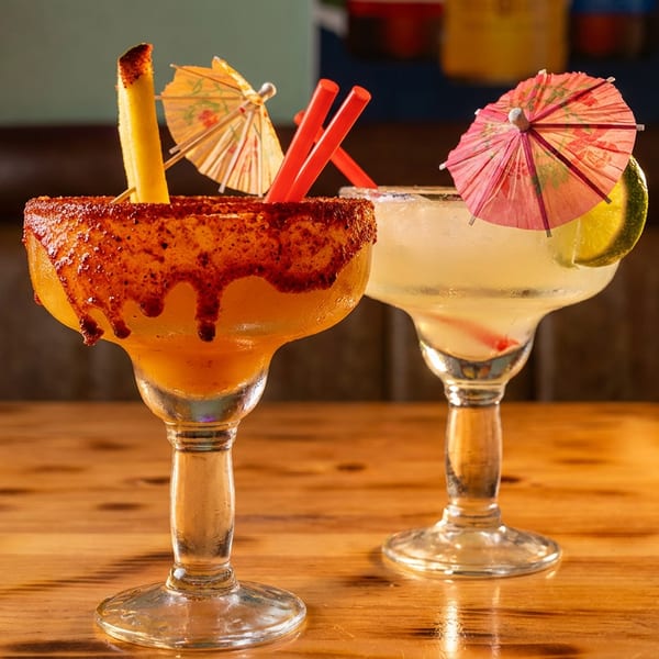 Two margaritas on a wooden table, one with a chili-lime rim and mango, the other with a lime.
