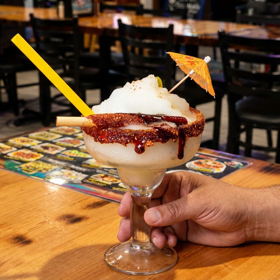 A hand holds a large frozen margarita with a chili-lime rim, tamarind candy straw, and umbrella.