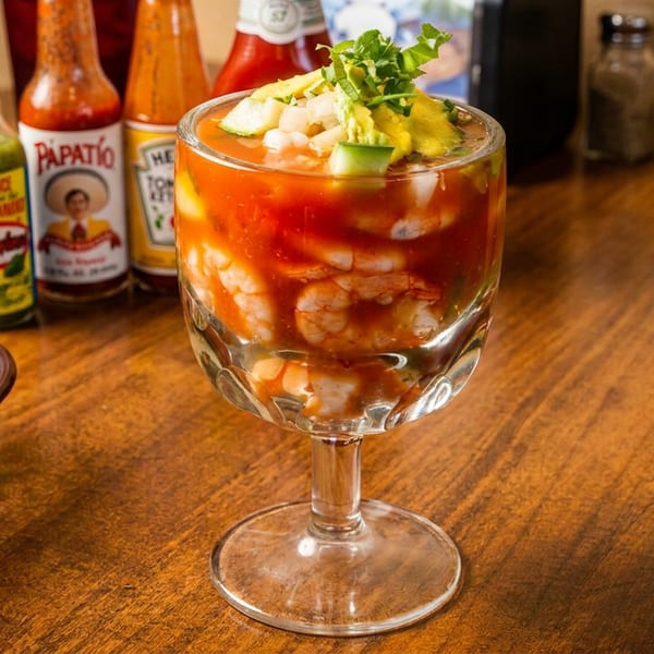 Shrimp cocktail in a goblet with avocado and cilantro, hot sauces and limes on a wooden table.
