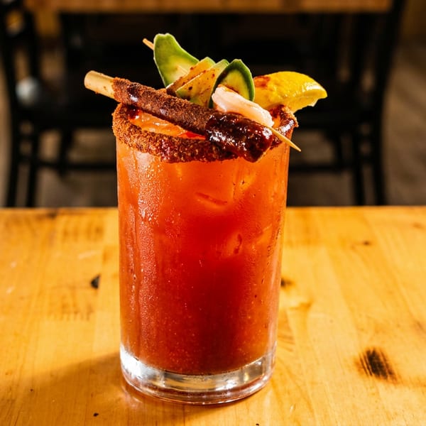 A vibrant red Michelada cocktail, garnished with avocado, shrimp, and a chili-dusted rim, on a wooden table.