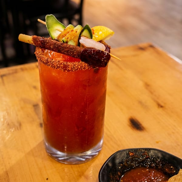 A tall glass of red michelada cocktail, garnished with avocado, shrimp, lemon, and a tamarind candy straw.