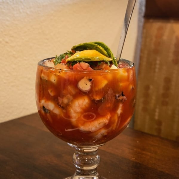A large glass of shrimp and octopus cocktail with avocado slices and a spoon on a wooden table.