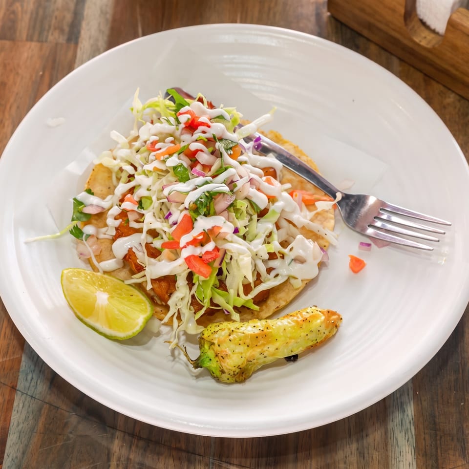 A tostada topped with cabbage, pico de gallo, and crema, served with a lime wedge and roasted pepper.