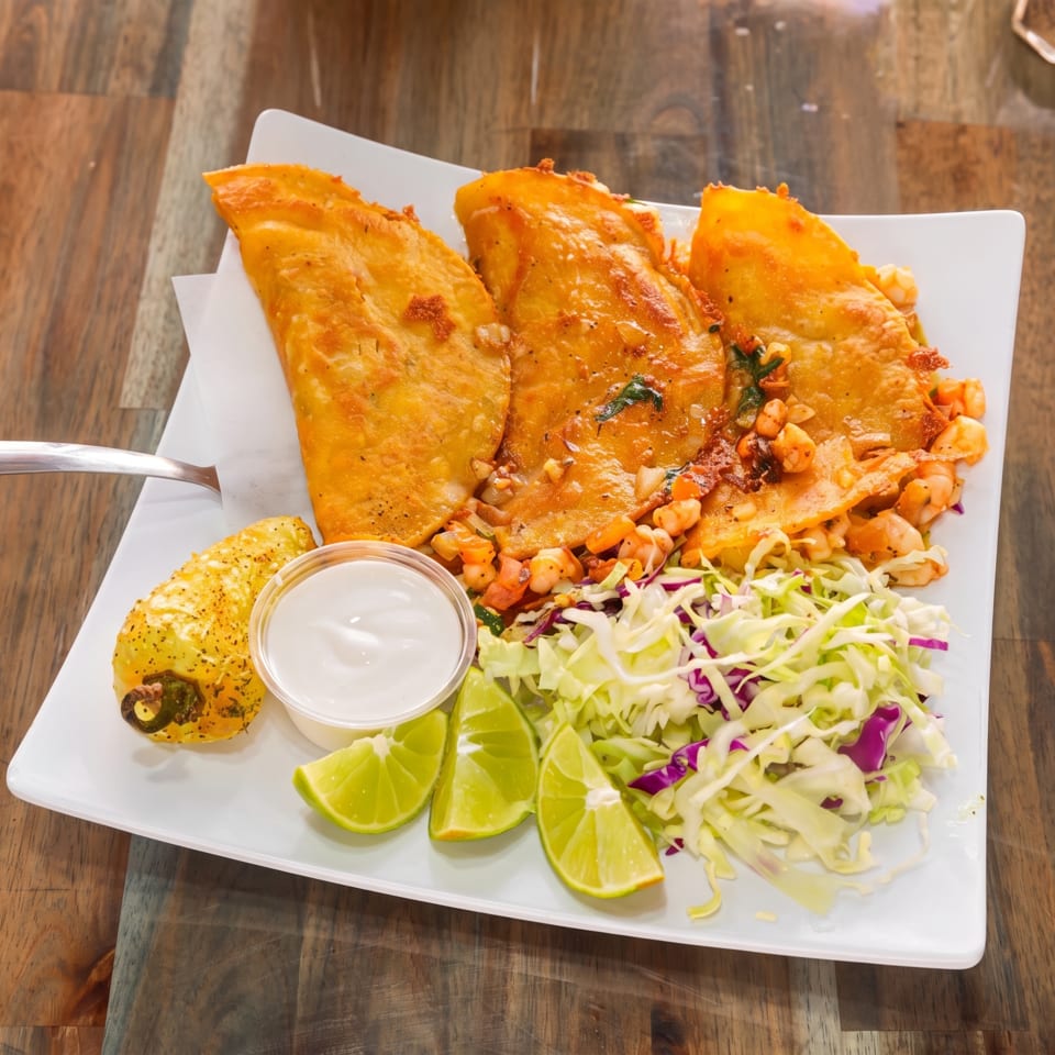Three golden-fried tacos with shrimp, cabbage, lime wedges, and a side of crema on a white plate.