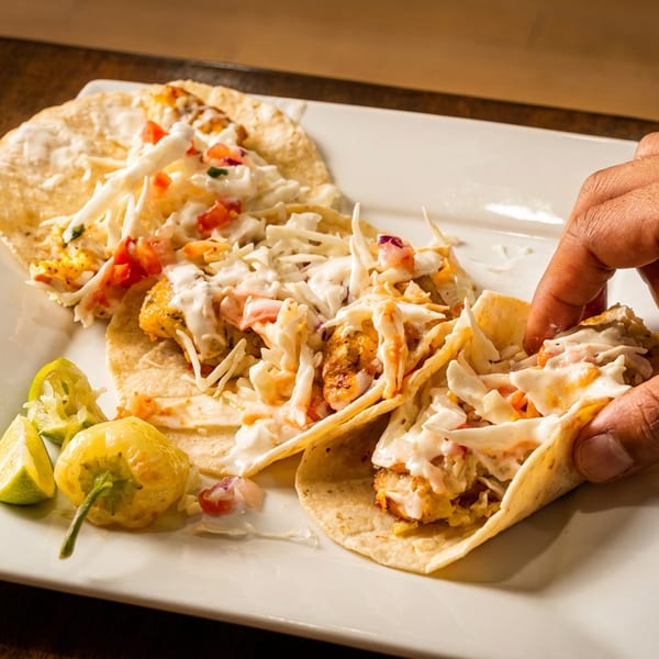 A hand reaching for a fish taco with cabbage, pico de gallo, and sauce on a white plate.